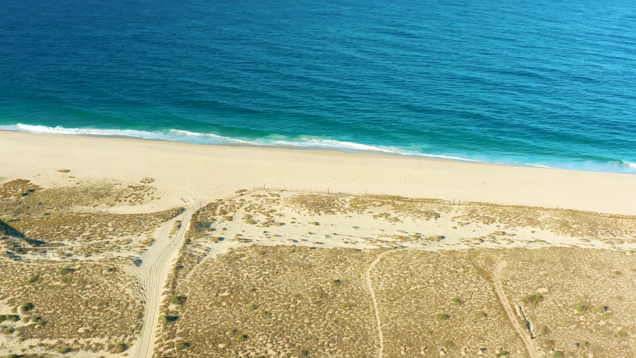 At 0:02, blue water and a narrow band of white surf sit above the pale Todos Santos beach, while dune scrub and winding sandy paths fill the lower frame with no visible people.