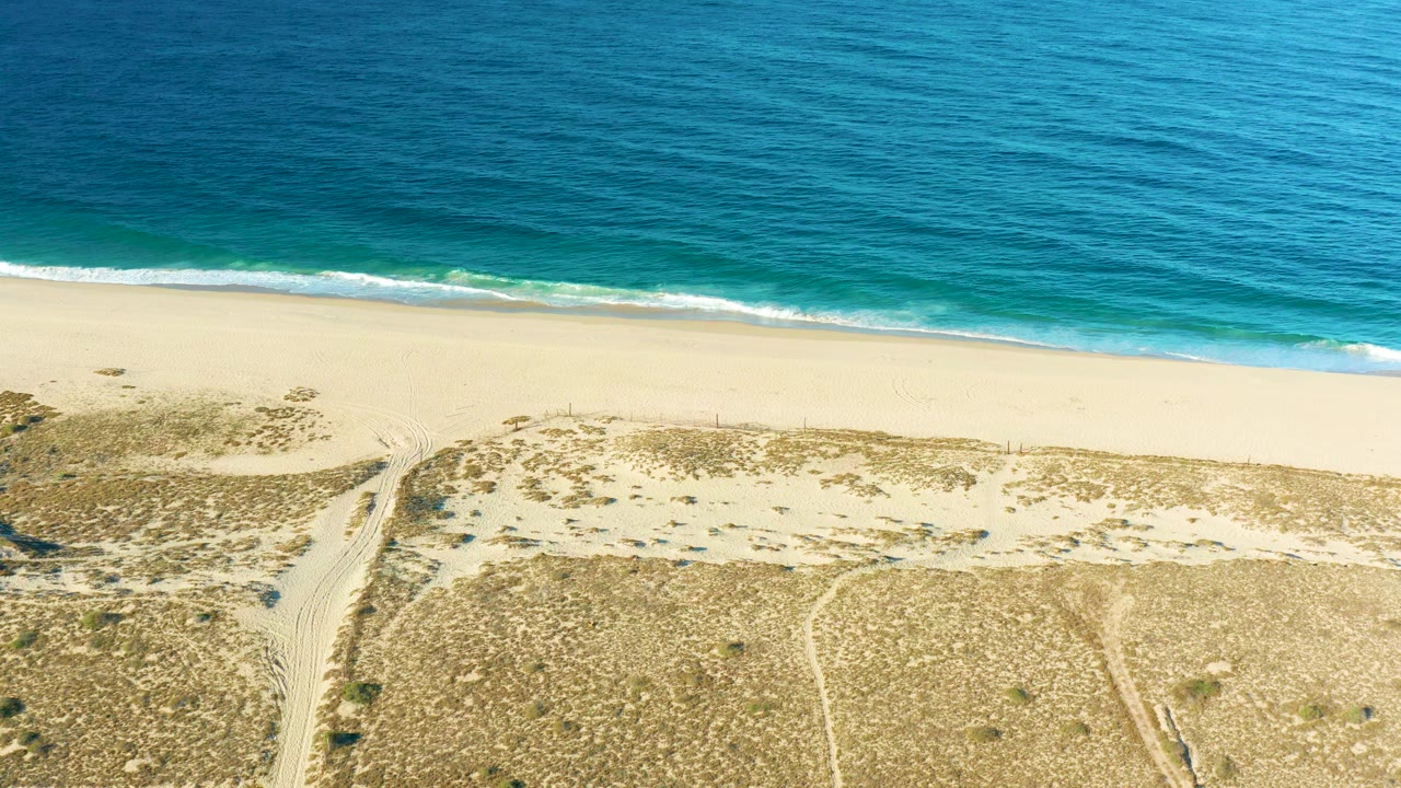 At 0:03, the foamy shoreline cuts across the frame below the blue ocean, with pale beach, low dunes, and a sandy access path visible in Todos Santos and no visible people.