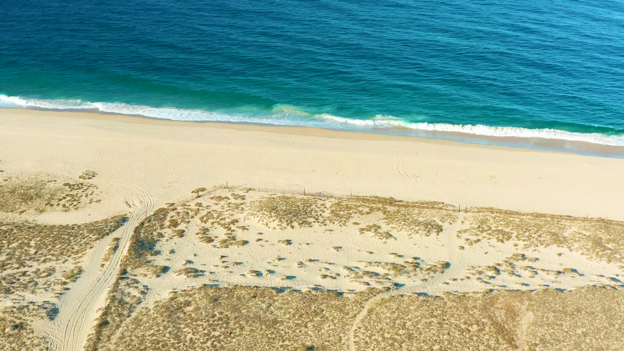 Aerial frame over Todos Santos showing turquoise nearshore water, a thin white wave line, pale beach, dune vegetation, and no visible people.