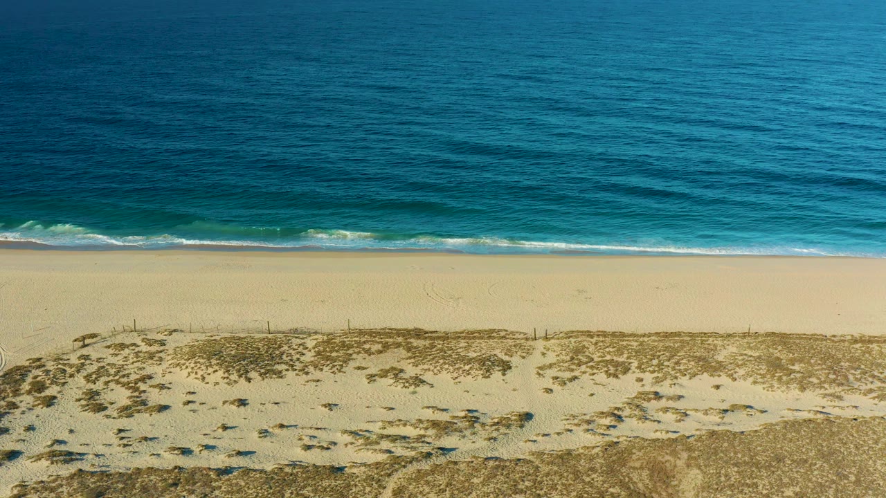 The Todos Santos shoreline remains centered with a narrow white wave edge between blue water and pale sand, and no visible people on the beach or dunes.