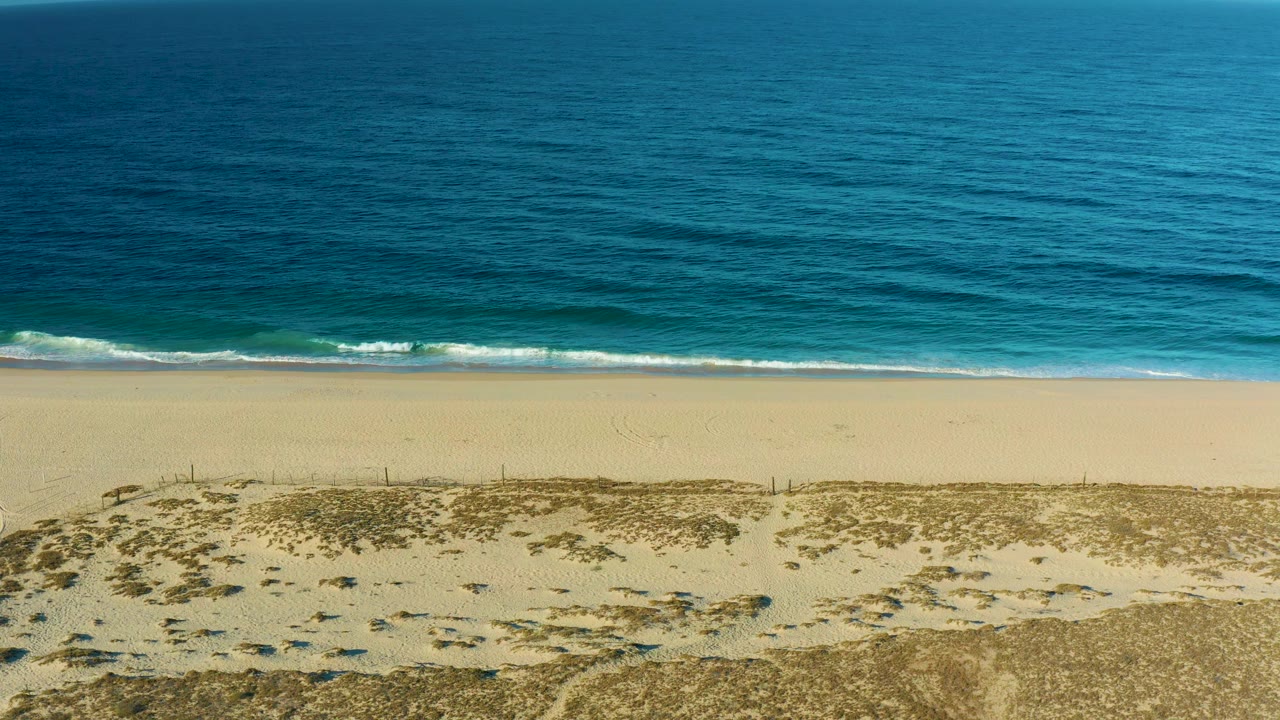 A slightly brighter frame shows the empty Todos Santos beach below deep blue water, with dune scrub across the lower foreground and no visible people.