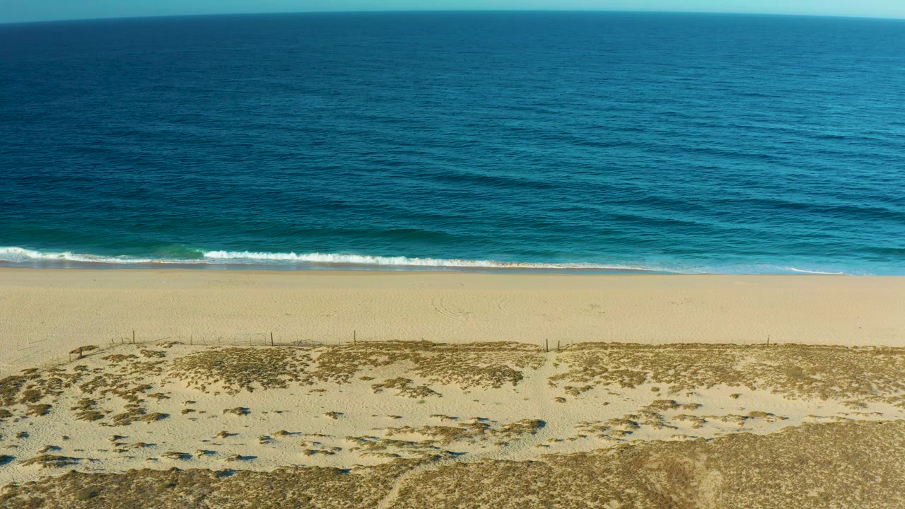 Midway through the clip, the Todos Santos coast appears as broad horizontal bands of scrub dunes, pale beach, thin surf, and open blue water, with no visible people.
