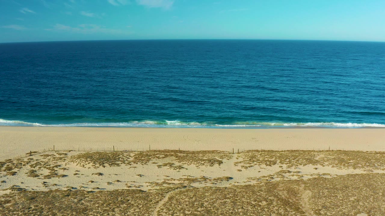 Todos Santos beach appears from above with sandy dunes in the foreground, a thin white surf line, deep blue ocean to the horizon, scattered clouds, and no visible people.