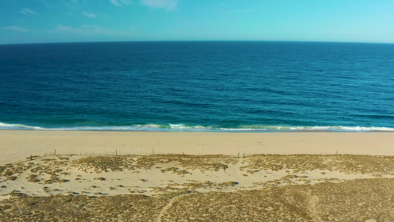 The second frame holds on the Todos Santos shoreline, with pale dunes at the bottom, a brighter band of sand, low waves near shore, blue ocean, light cloud patches, and no visible people.