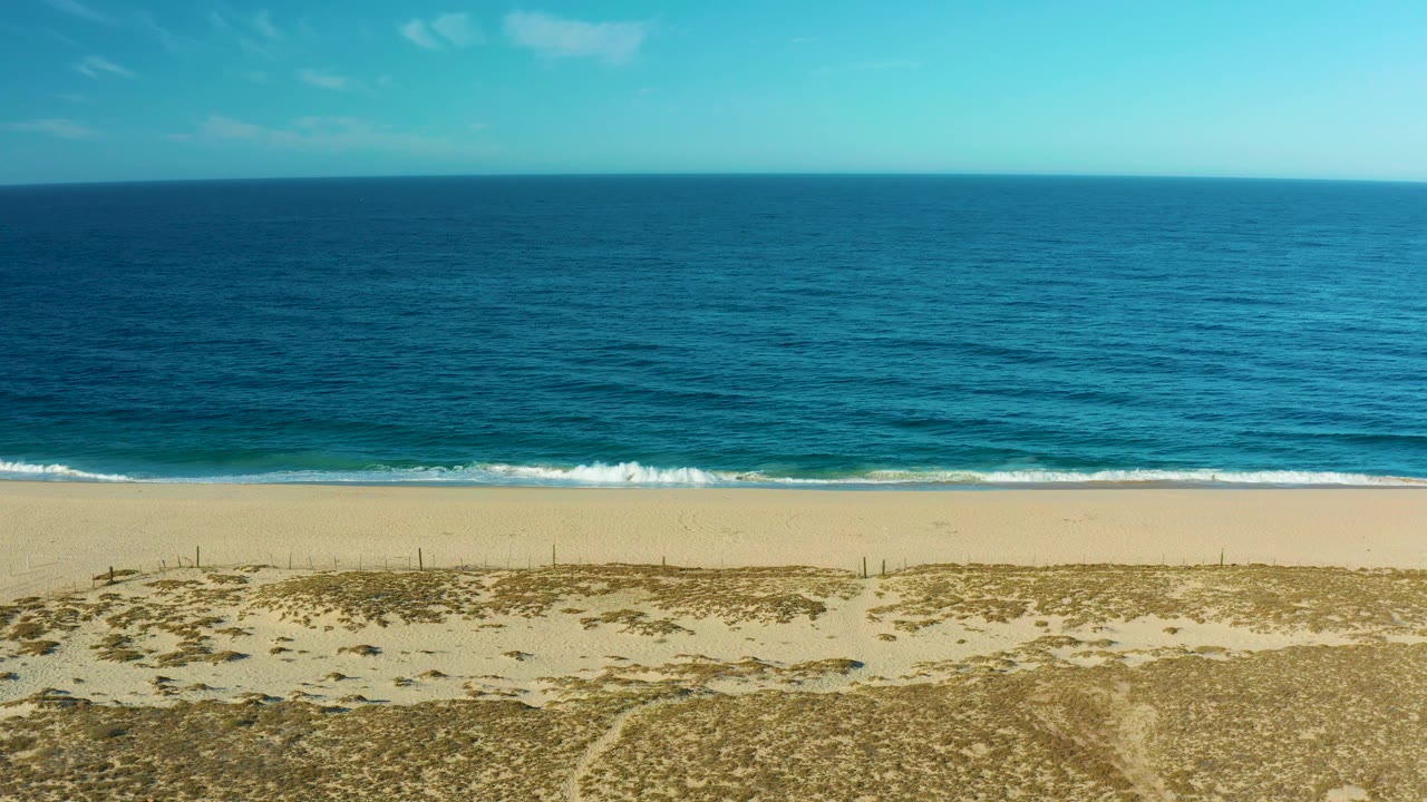 Frame three shows the Todos Santos coast with darker blue water across the upper frame, a narrow white wave edge, open sand, dry dune vegetation texture, and no visible people.
