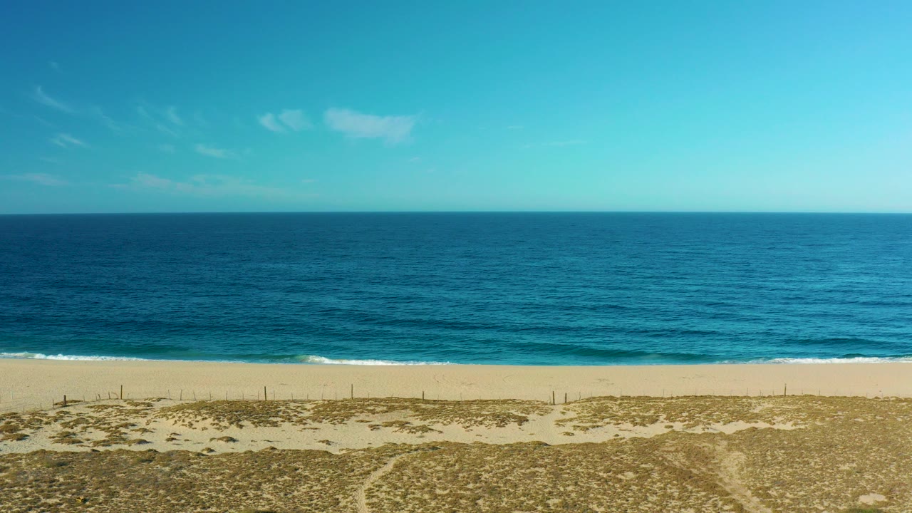 Wide beach, dunes, blue ocean, and clear sky.
