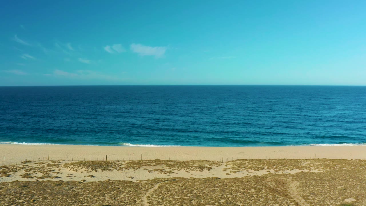 A Todos Santos shoreline frame shows a straight ocean horizon, gentle surf near the sand, dry dune vegetation in front, and no visible people.