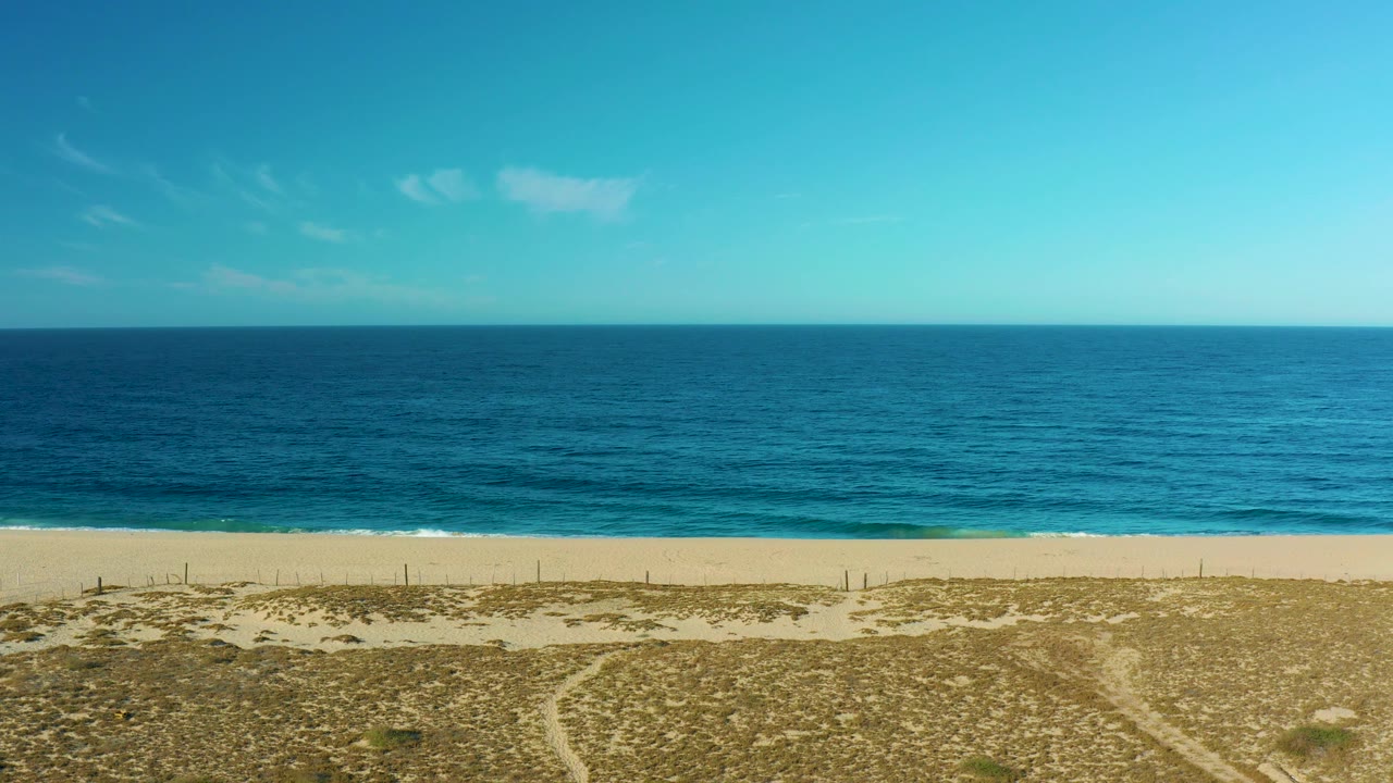 Mid-clip view of the Todos Santos beach shows empty sand, a band of breaking surf, deep blue water, pale cloud patches, and no visible people.