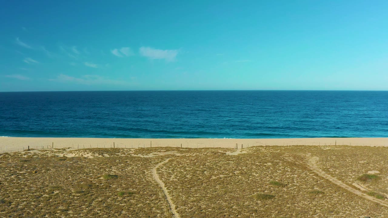 Early frame shows the Todos Santos shoreline with the horizon high across the image, a pale beach strip, low dune grass, branching sandy tracks, and no visible people.