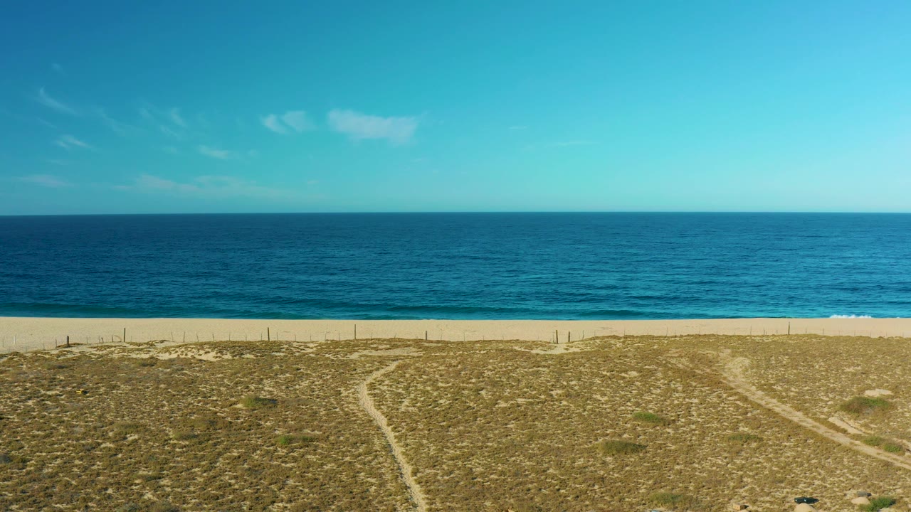 Aerial view over Todos Santos dunes where a curving sandy path cuts through dry grass toward the beach, with calm blue ocean behind it and no visible people.