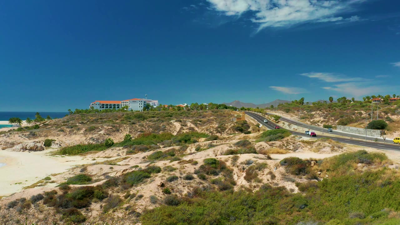 A second view of the Marquis Los Cabos coastal road shows cars spaced along the pavement, sandy hills and green scrub in the foreground, ocean to the left, and no visible people.