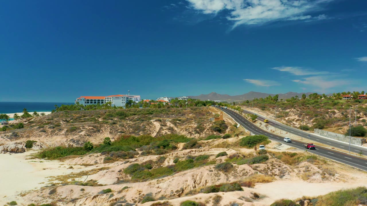 Wide aerial-style frame of the Marquis Los Cabos coastal highway with vehicles shifted along the lanes, pale sand and shrubs in the foreground, ocean at left, and no visible people.