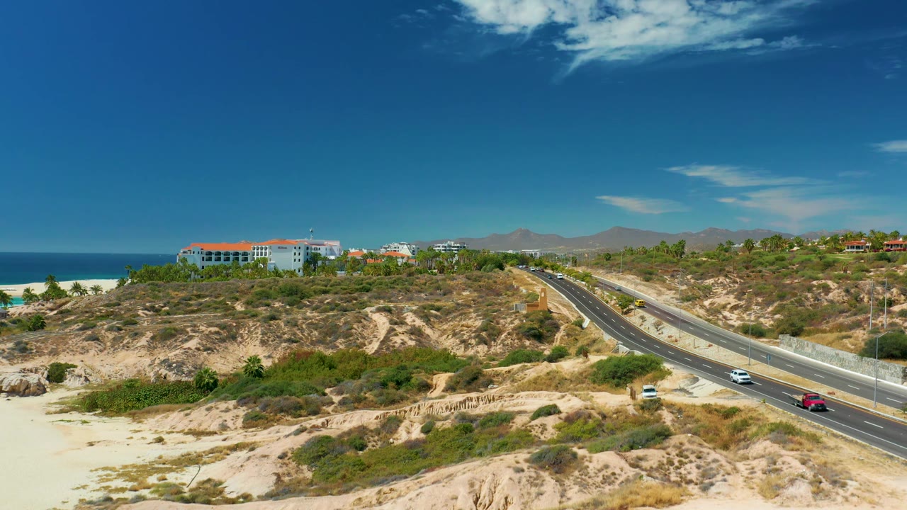 The road angles through the right side of the Marquis Los Cabos frame as cars travel past scrub-covered sandy hills, with ocean, beach, and hilltop buildings visible and no visible people.