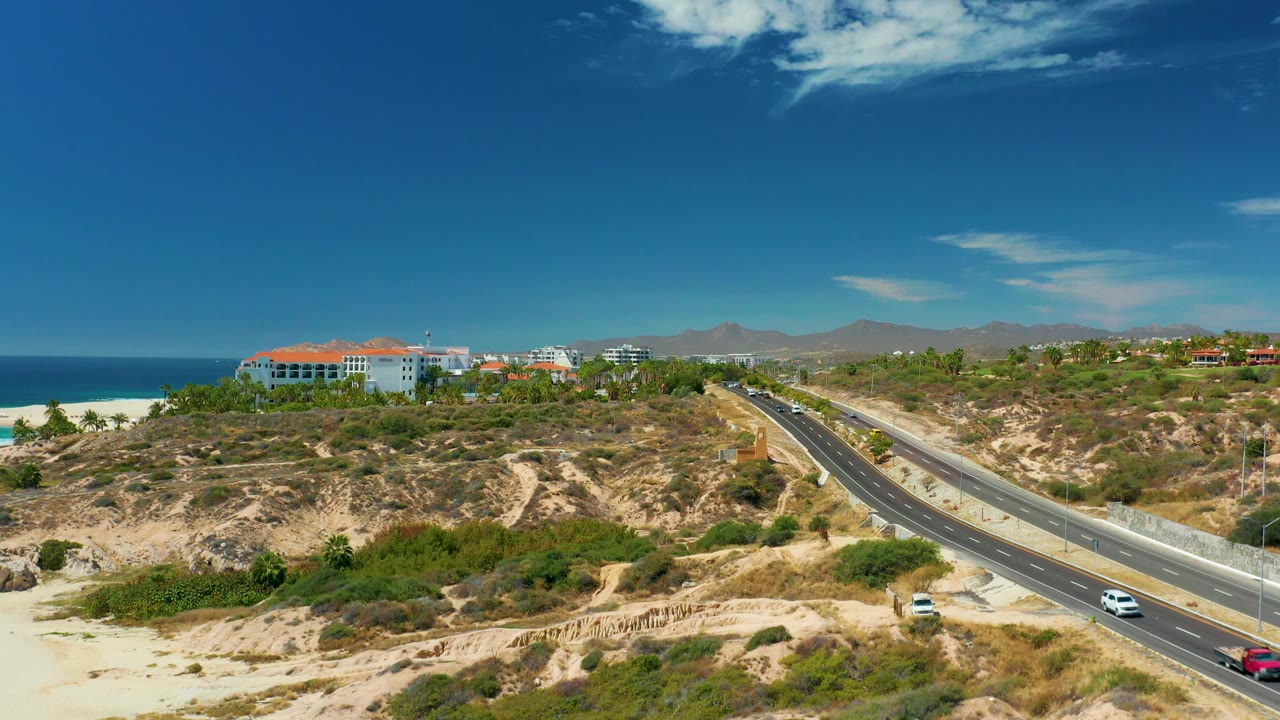 A white car and red car are visible on the Marquis Los Cabos highway, with dry foreground slopes, sparse shrubs, ocean to the left, buildings above the beach, and no visible people.