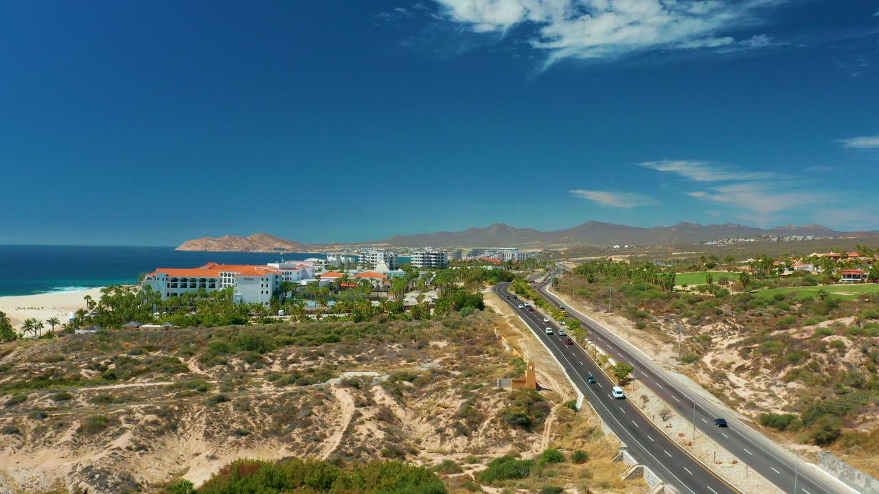 Aerial view of a coastal road beside beach, ocean, buildings, hills, and cars.