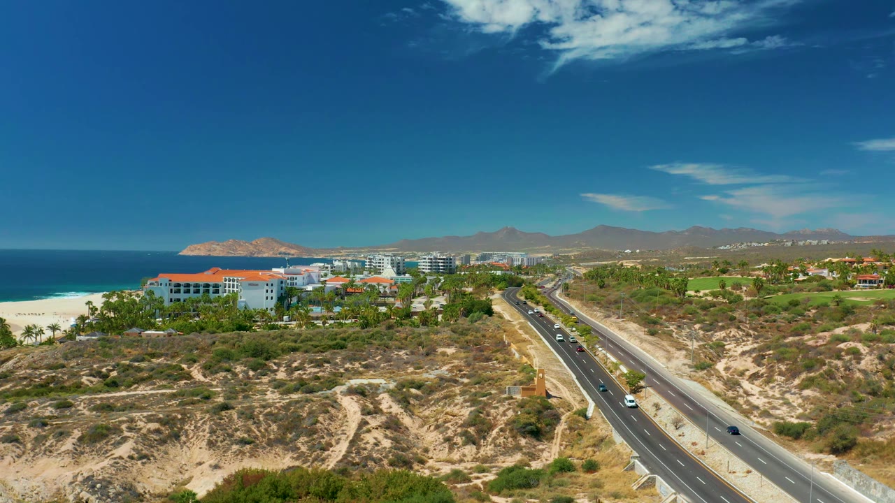 Aerial view at 0:01 over Marquis Los Cabos, Mexico, with cars spread along the diagonal road, white beach and ocean on the left, buildings near the shore, hills beyond, and no visible people.