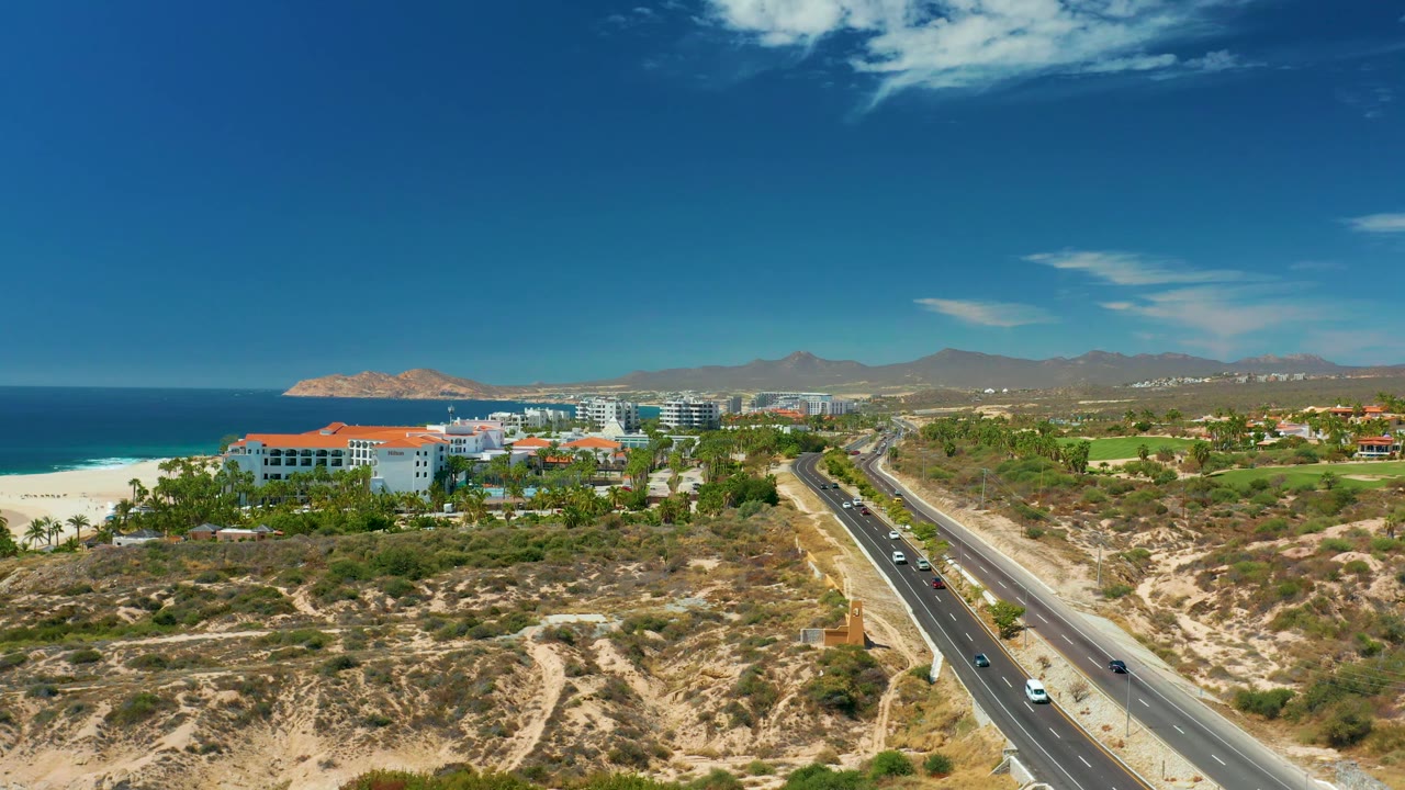 Aerial frame at 0:02 showing Marquis Los Cabos coastal traffic, with the road edging closer to the right side, beachfront buildings centered, ocean and sandy beach to the left, hills in back, and no visible people.