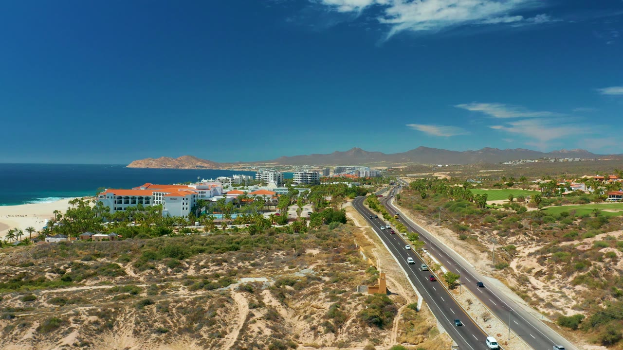 Aerial view at 0:03 over Marquis Los Cabos, Mexico, with more beachfront buildings visible near the beach, several cars on the diagonal road, scrub vegetation in the foreground, hills beyond, and no visible people.