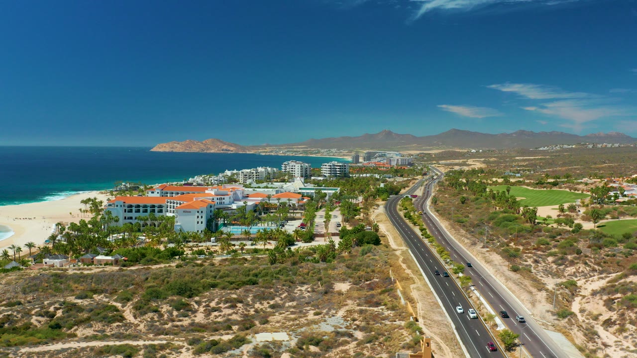 Wide aerial frame over Marquis Los Cabos showing white resort buildings with orange roofs, sandy beach, blue ocean, parallel highway, dry hills, distant mountains, and no visible people.