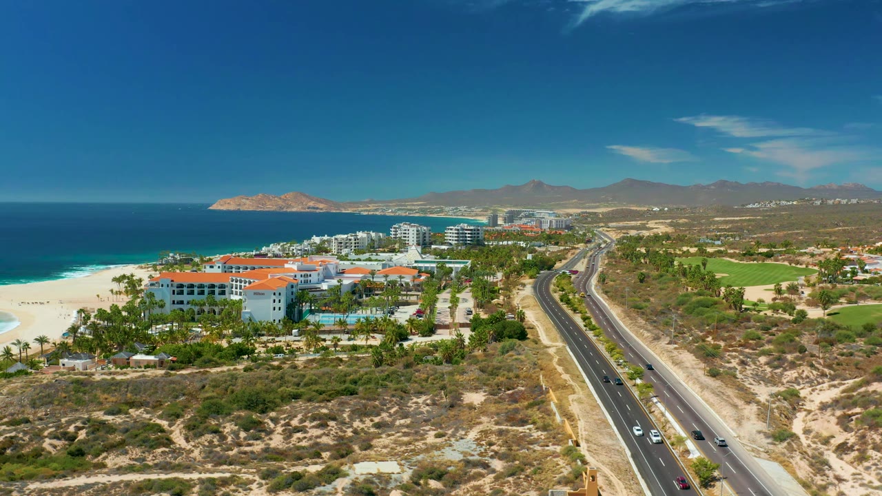Aerial view at Marquis Los Cabos with the resort complex left of the highway, blue ocean and pale beach to the side, cars on the road, mountains on the horizon, and no visible people.