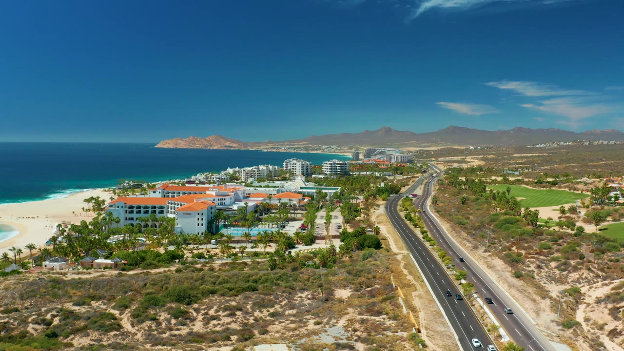High aerial frame of Marquis Los Cabos with white hotel blocks, orange rooflines, beach and turquoise surf, a divided highway carrying vehicles, dry land, and no visible people.