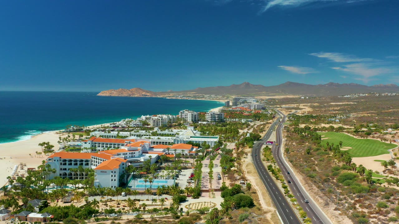 Aerial view of beach resorts, ocean, coastal road, fairway, and mountains.