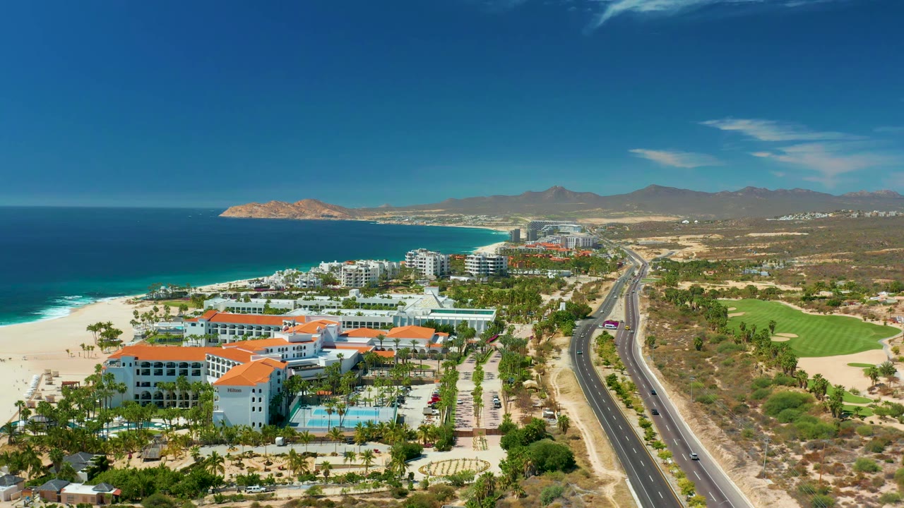Marquis Los Cabos aerial frame one second in, showing more of the beachfront hotels and red roofs below the camera, ocean and beach to the left, a divided road with cars, fairway grass to the right, distant mountains, and no visible people.