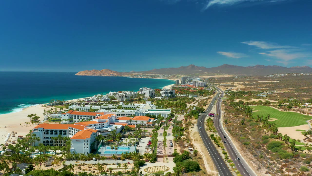 Marquis Los Cabos aerial frame at 0:02, with the beach and blue water running along the left edge, resort blocks and palms in the lower frame, cars on the road, green fairway land at right, low mountains beyond, and no visible people.