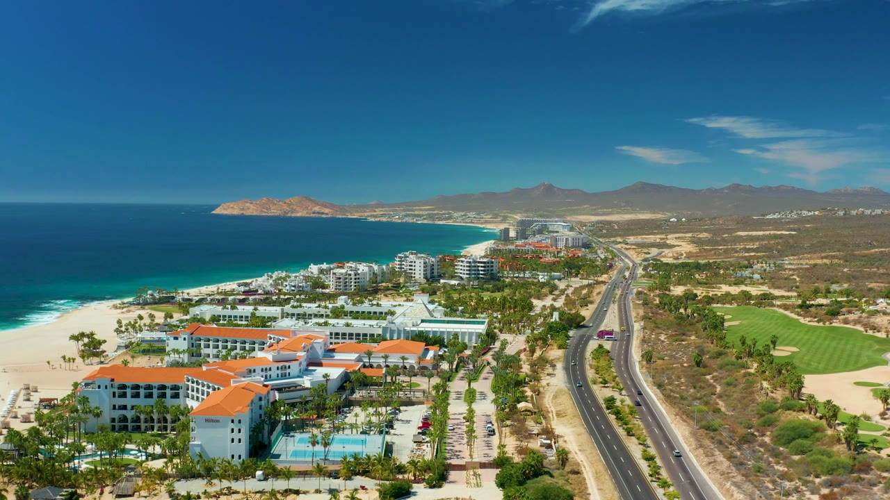 Mid-clip aerial over Marquis Los Cabos, Mexico, where the ocean and sandy shoreline fill the left side, hotel buildings and palms sit below, the highway curves through the center, green fairway sections lie to the right, and no visible people appear.