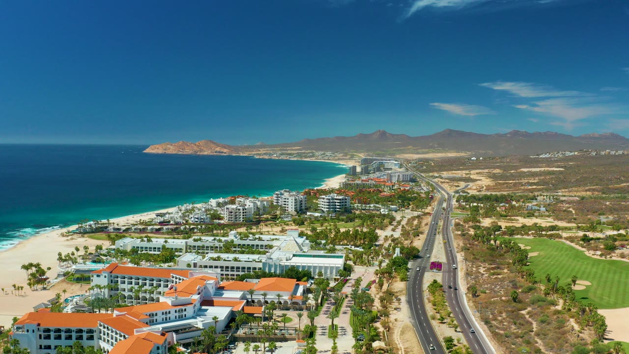 Wide aerial frame over Marquis Los Cabos, Mexico, with blue ocean and surf on the left, sandy beach and white buildings below, a straight road to the right, mountains on the horizon, and no visible people.