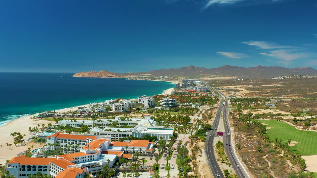 Aerial view at Marquis Los Cabos, Mexico, showing the ocean band, pale beach, clustered coastal buildings, a road running inland-side of the shore, green golf course areas, distant mountains, and no visible people.