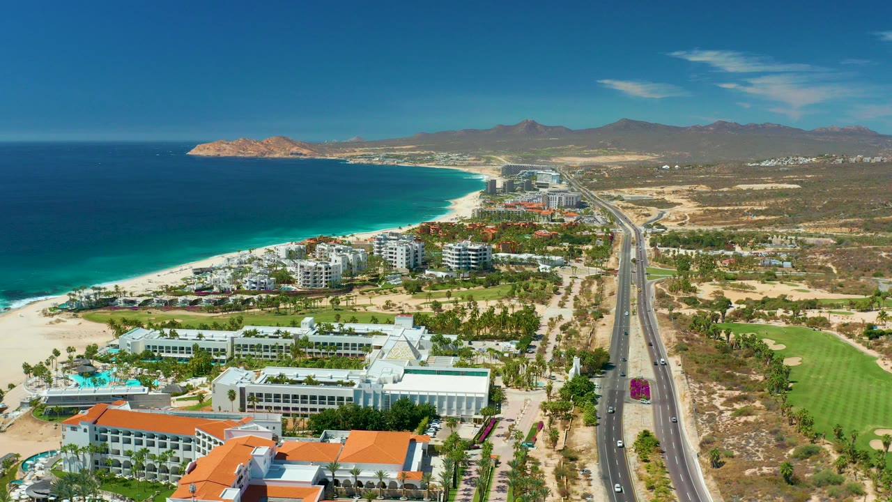 Wide aerial frame over Marquis Los Cabos, Mexico, with turquoise ocean and white beach on the left, resort buildings below, a highway through the center, golf fairways on the right, mountains in the distance, and no visible people.