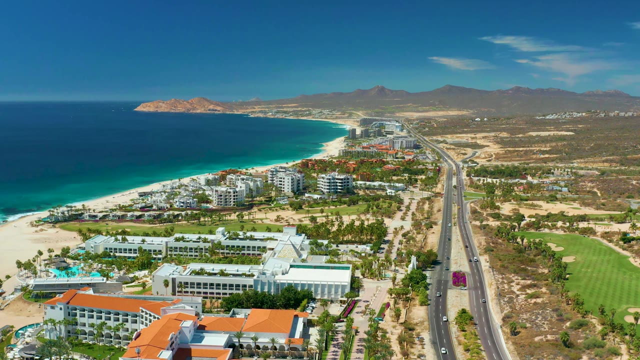 Elevated view along the Marquis Los Cabos coastline, showing the beach and blue ocean to the left, white resort buildings and pools in the foreground, the highway running away from camera, green fairways to the right, and no visible people.