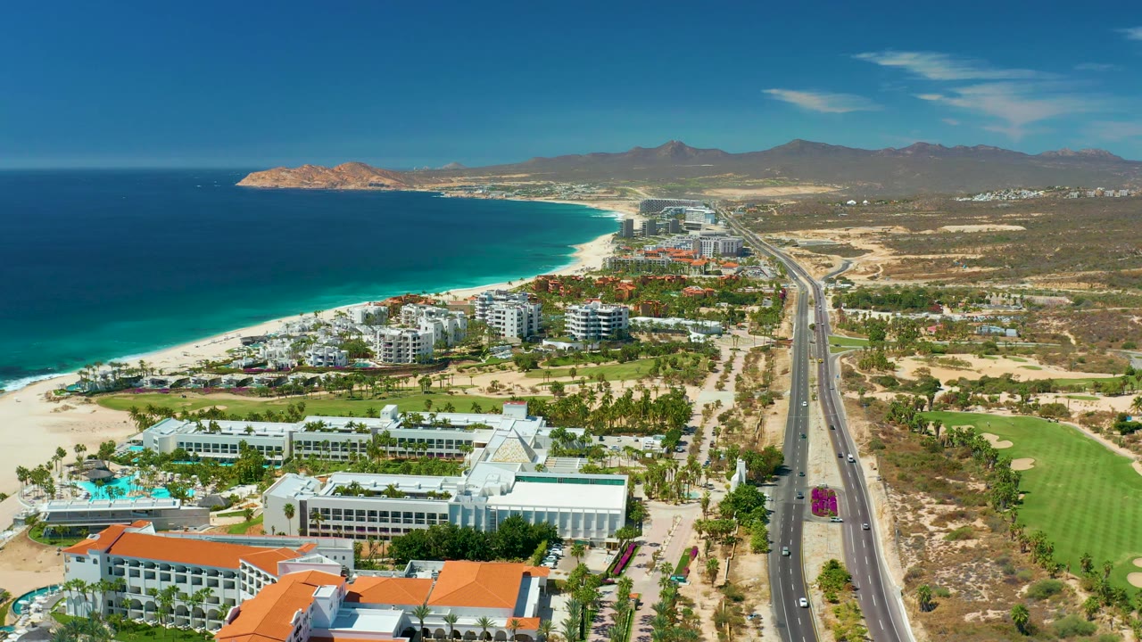 Drone-height frame over Marquis Los Cabos with a broad band of ocean, a pale sandy shoreline, beachfront hotel buildings, a straight highway with a red car near the lower road, golf greens to the right, and no visible people.