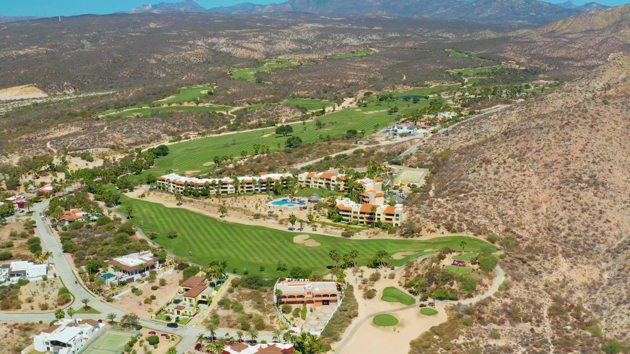 Aerial view one second into the Marquis Los Cabos clip with the golf course centered, a road along the left side, resort-style buildings below the hills, and no visible people.
