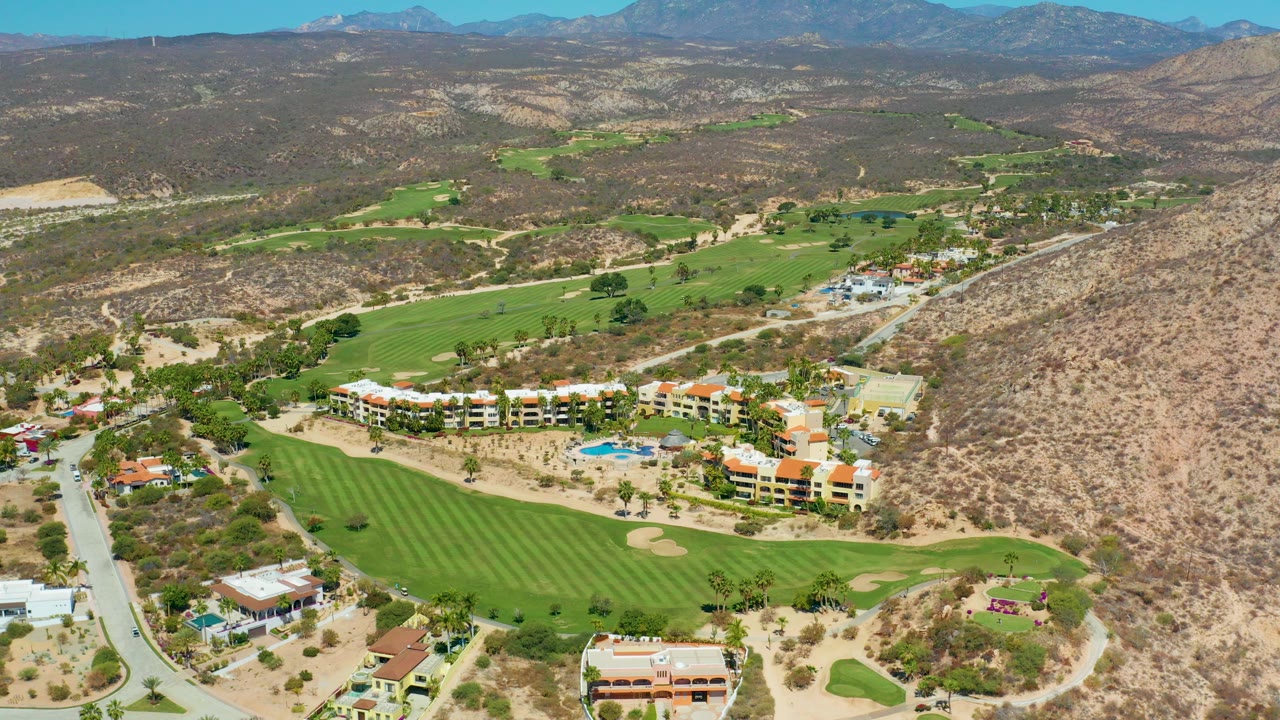 Aerial frame over Marquis Los Cabos, Mexico showing green fairways, sand bunkers, low buildings, a blue pool, roads, dry hills, distant mountains, and no visible people.