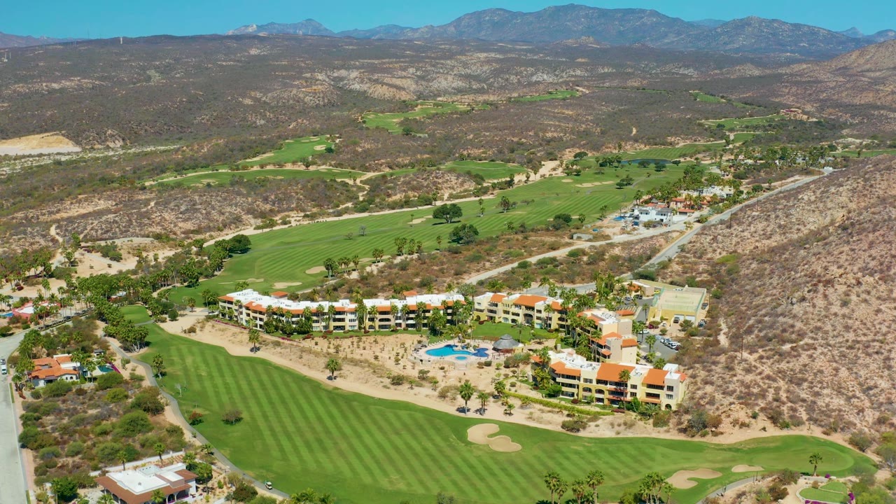 Early frame from the aerial pass at Marquis Los Cabos, Mexico, with the resort complex centered lower in frame, fairways to the left, dry hills to the right, and no visible people.