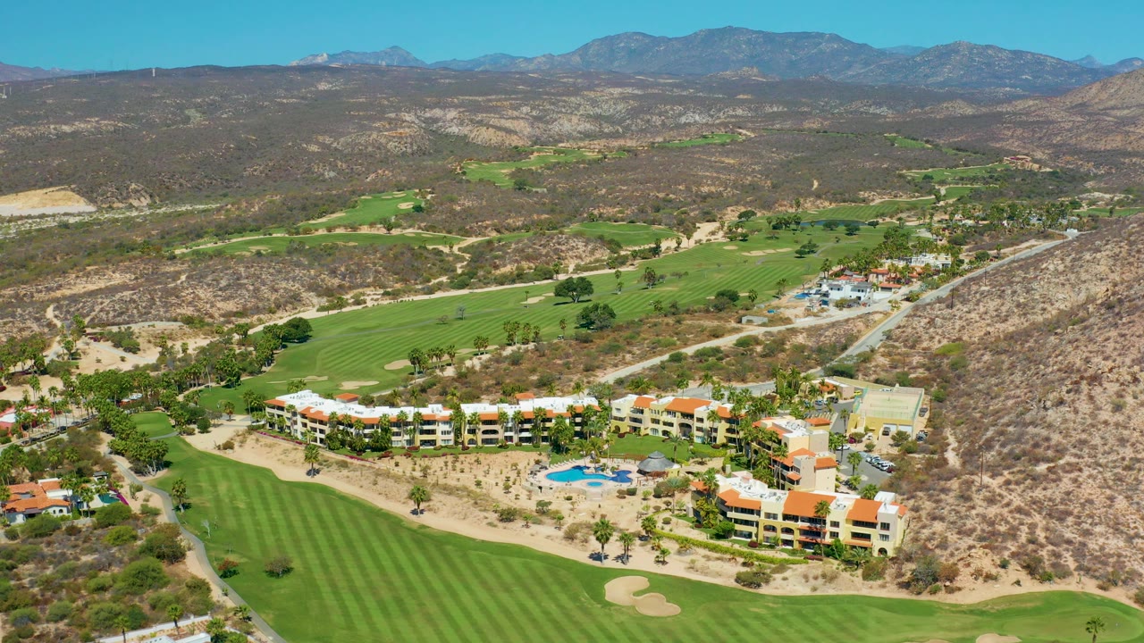 Mid-clip aerial frame over Marquis Los Cabos, Mexico, with resort buildings along the lower edge, bright fairways stretching into the valley, dry hills around them, and no visible people.
