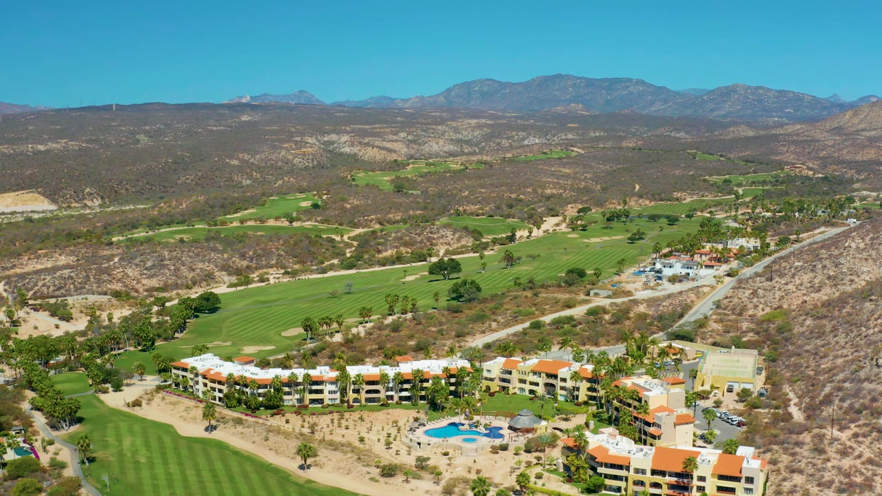 Marquis Los Cabos aerial view with the resort complex lower in the frame, golf greens stretching across the valley, a road cutting through dry terrain, distant mountains, and no visible people.