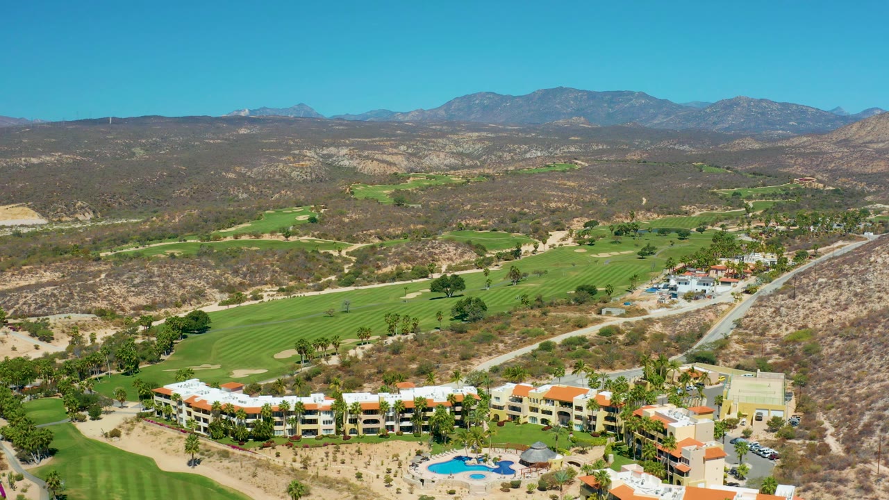 Mid-clip aerial frame at Marquis Los Cabos with resort roofs partly along the bottom, a road running into the valley, golf fairways on both sides, dry brush-covered hills, mountains, and no visible people.