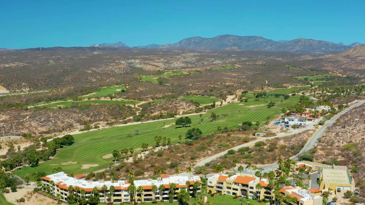 Aerial frame at Marquis Los Cabos, Mexico, with the golf course spread across the center, resort buildings along the lower edge, a road on the right, dry hills, mountains, and no visible people.