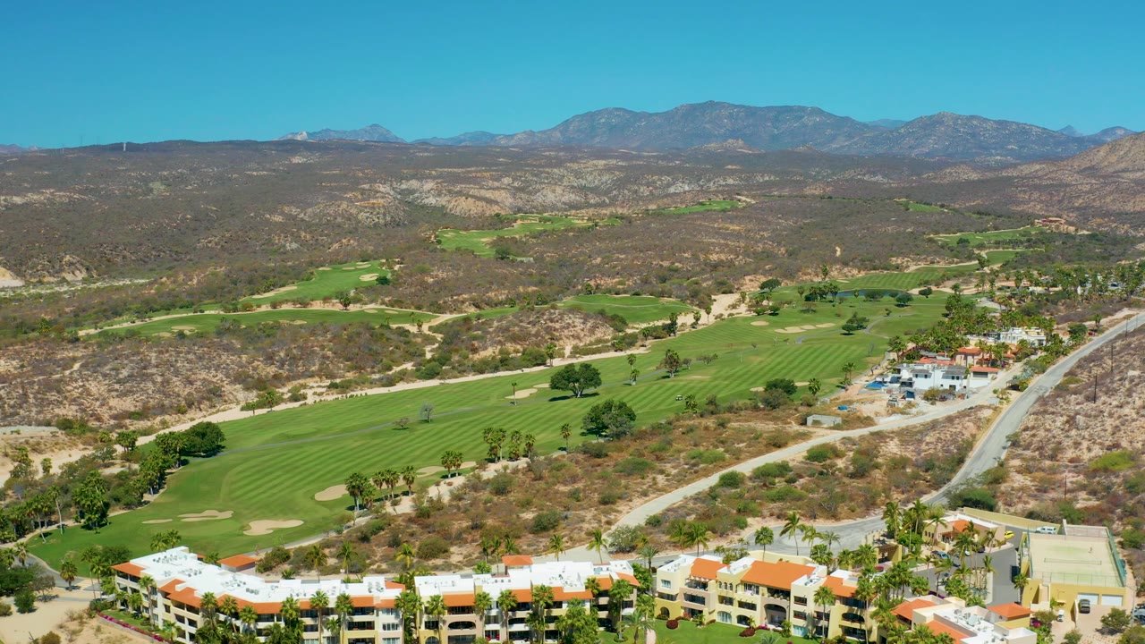 High aerial view at Marquis Los Cabos, Mexico, with bright green fairways in the lower half, pale resort buildings near the bottom, desert hills across the middle, distant mountains, and no visible people.