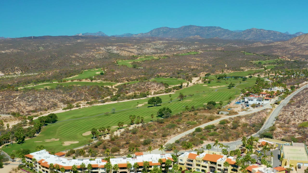 Aerial view at Marquis Los Cabos, Mexico, showing a curving road beside the course, clustered resort buildings at the lower right, palm trees, fairways, dry hills, mountains, and no visible people.