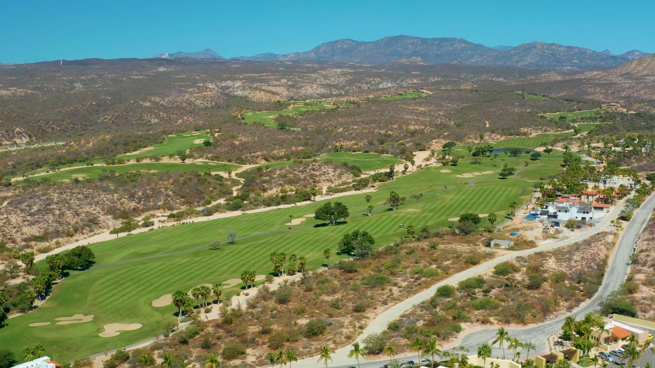 Aerial view at Marquis Los Cabos, Mexico showing green golf fairways, sand traps, dry hills, a road on the right, distant mountains, clear blue sky, and no visible people.