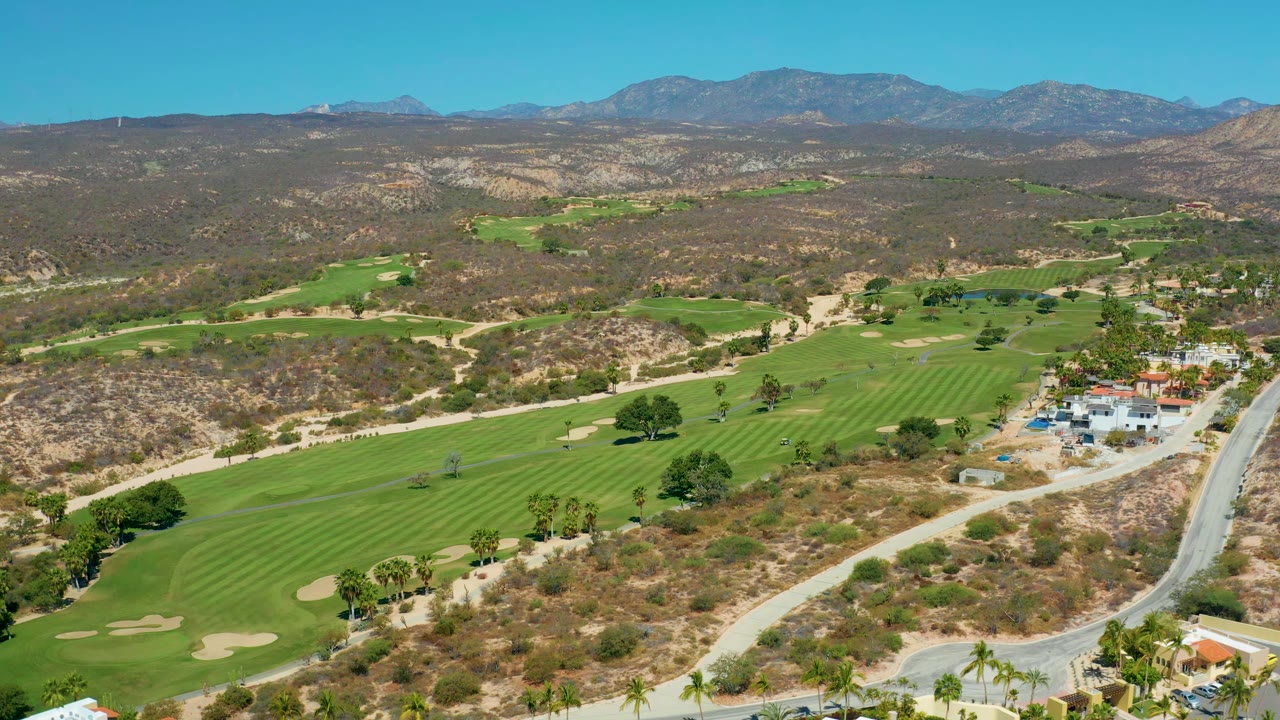Marquis Los Cabos aerial frame with striped green fairways, pale sand traps, scattered palms, dry brush-covered hills, distant mountains, clear sky, and no visible people.