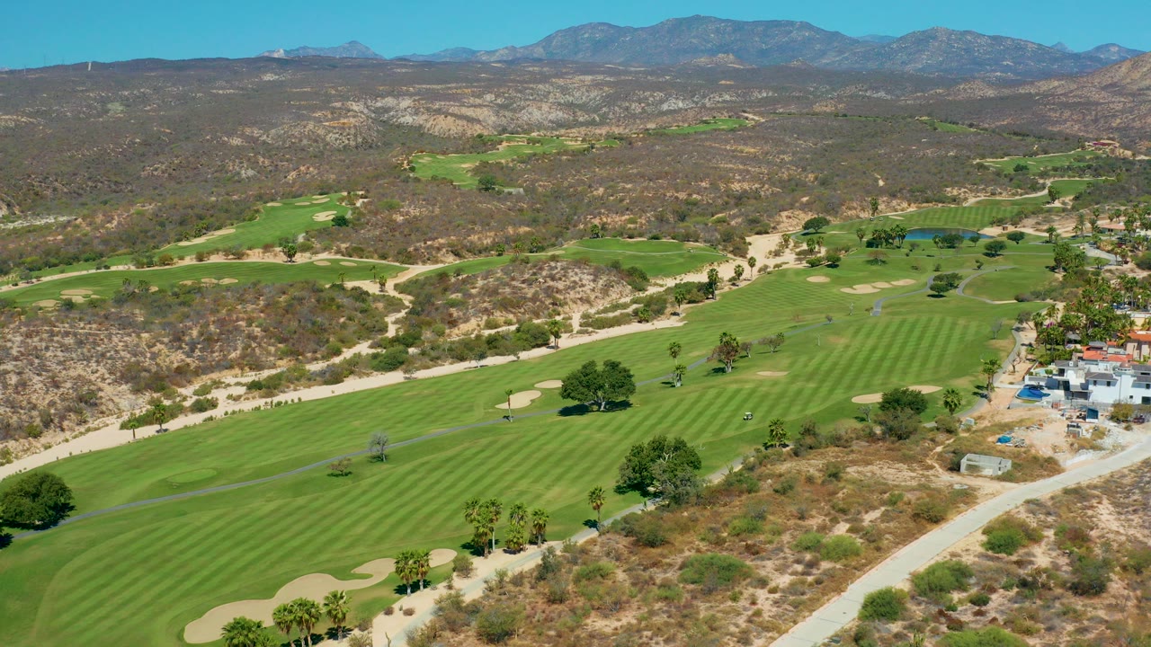 Aerial frame over Marquis Los Cabos, Mexico showing striped green fairways, sand traps, dry hillside terrain, distant mountains, and no visible people.
