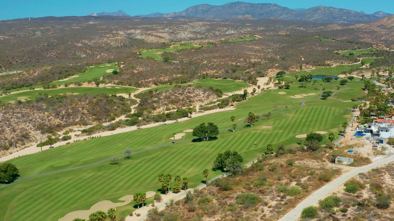 High aerial frame over Marquis Los Cabos, Mexico showing a diagonal road, striped fairways, tan bunkers, scrub-covered hills, mountains, and no visible people.