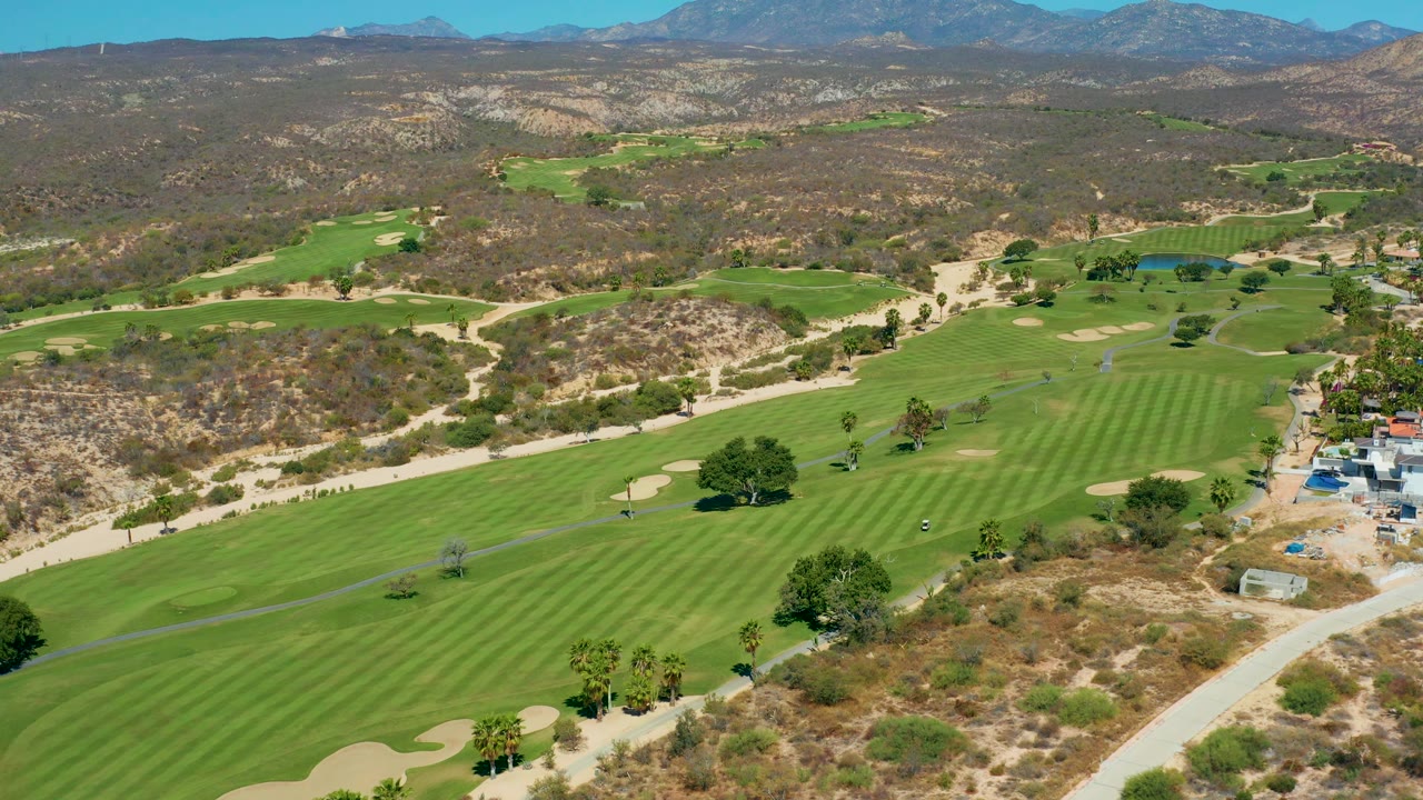 Wide aerial view of golf fairways and dry hills with buildings at the right edge.