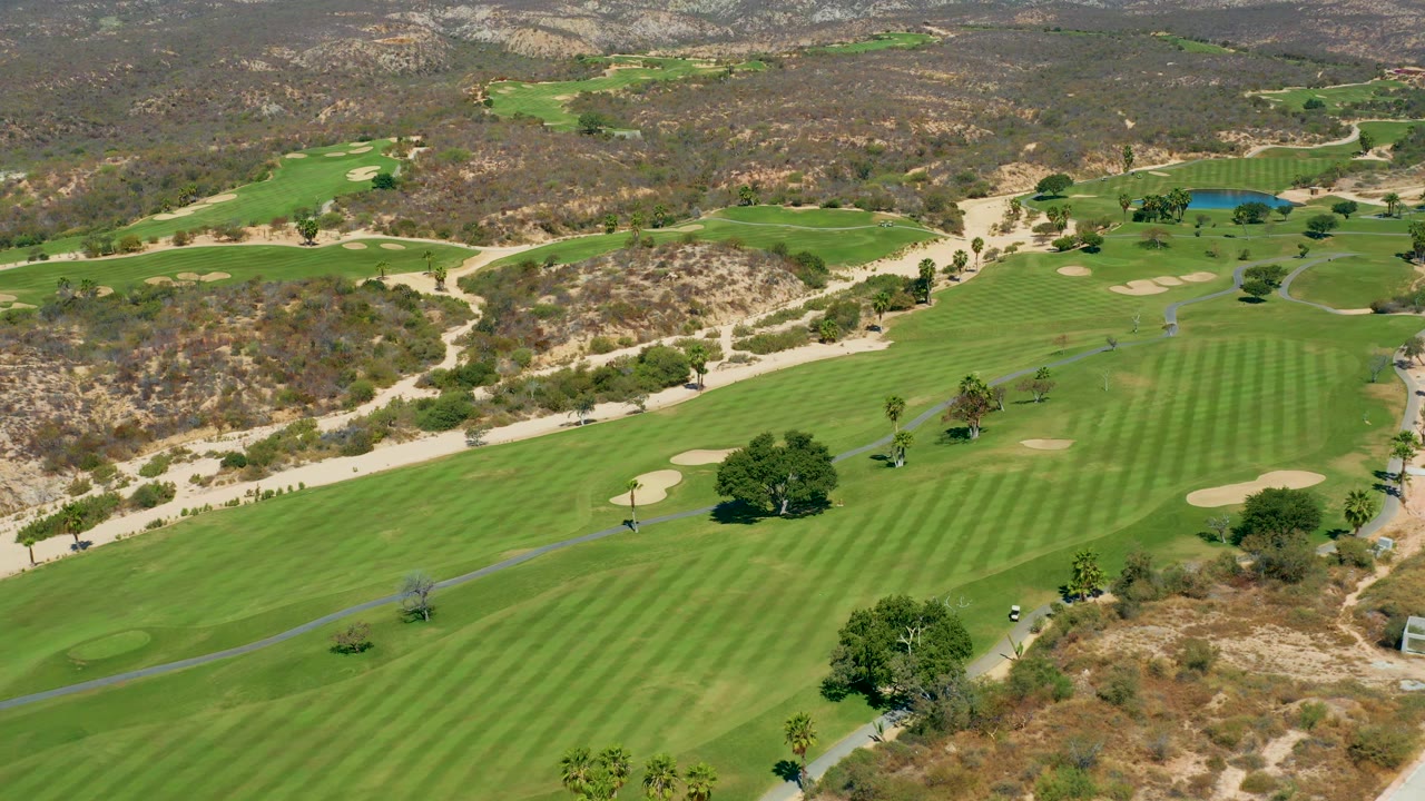 Aerial view across the Marquis Los Cabos course with a broad striped fairway in the center, dry hillside ground beyond it, and no visible people.
