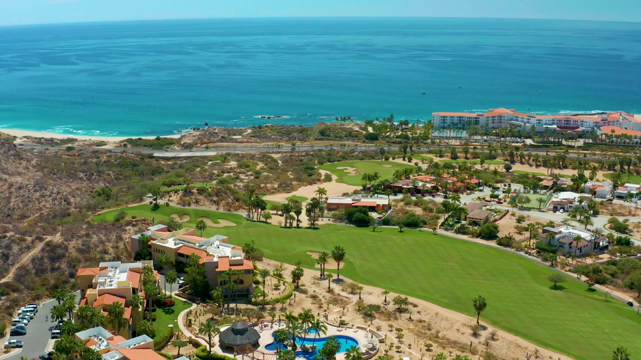 Wide aerial view of turquoise ocean, sandy shoreline, golf fairway, palms, and buildings.